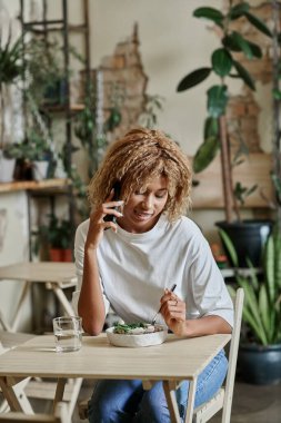 Telli mutlu Afrikalı Amerikalı kız akıllı telefonla konuşuyor ve kafede taze vegan salatası yiyor.