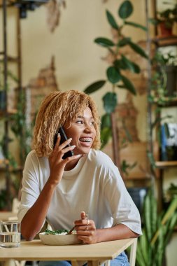 Telli neşeli Afrikalı Amerikalı kız akıllı telefondan konuşuyor ve kafede taze vegan salatası yiyor.