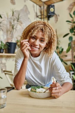 cheerful dark skinned girl with braces holding fork near fresh salad bowl in modern vegan cafe clipart