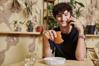 Mutlu ve kıvırcık genç adam elinde bitki bazlı tofu burgeri tutuyor ve vegan kafede akıllı telefondan konuşuyor.