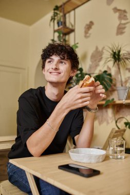 Mutlu ve kıvırcık genç adam organik tofu burgeri tutuyor ve bitki dolu vegan kafede gülümsüyor.