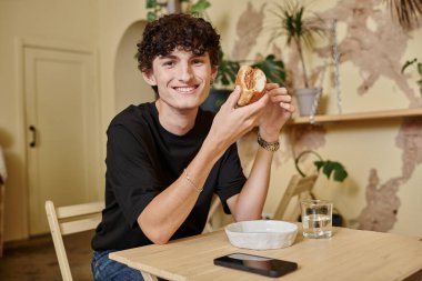 Neşeli ve kıvırcık genç adam organik tofu burgeri tutuyor ve sebze dolu kafede gülümsüyor.
