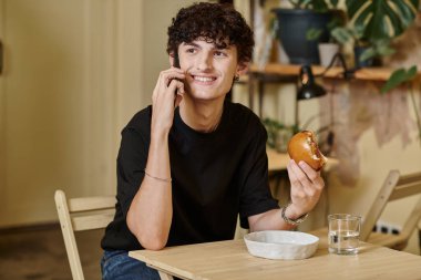 Mutlu ve kıvırcık genç adam elinde bitki bazlı tofu burgeri tutuyor ve vegan kafede akıllı telefondan konuşuyor.