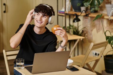 Kablosuz kulaklıklı mutlu adam dizüstü bilgisayar kullanıyor ve vejetaryen kafede tofu burgerinin tadını çıkarıyor.