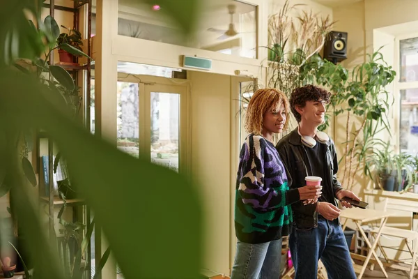 joyful and diverse couple in casual attire walking with plant based cappuccino in vegan cafe