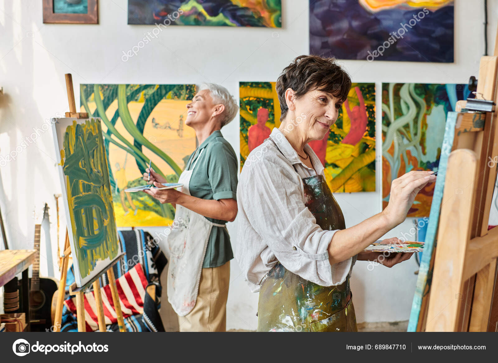 Скачать - Веселые стильные зрелые женщины в фартуках, указывающие на мольберты в художественной мастерской, дружба и творчество — стоковое изображение Веселые Стильные Зрелые Женщины Фартуках Указывающие Мольберты Художественной Мастерской Дружба — стоковое фото