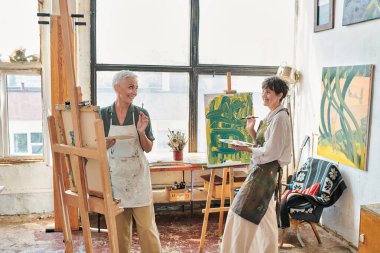 happy middle aged women in aprons painting on easels in modern art workshop, creative leisure clipart