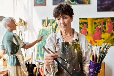 Sanat stüdyosunda resim yapan bir kadın arkadaşın yanında boya fırçası seçen yaratıcı olgun kadın ressam.