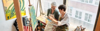 smiling middle aged women mixing colors on palettes near easels in art studio, horizontal banner clipart