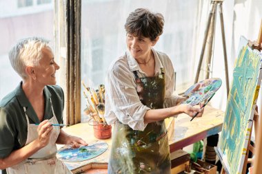 Sanat atölyesinde yüksek lisans yaparken paletleri olan mutlu olgun kadın sanatçılar resim sehpasının yanında konuşuyorlar.