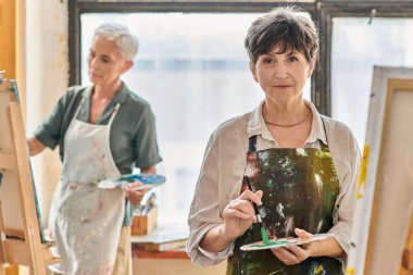 Sanat atölyesinde yüksek lisans yaparken elinde renk paleti olan ve kameraya bakan yaratıcı olgun bir kadın.