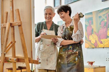 cheerful mature woman mixing paint on palette near smiling female artist, master class in art studio clipart