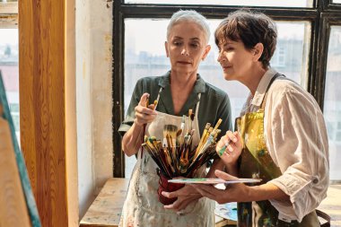 Yetenekli kadın sanatçı, sanat atölyesinde yüksek lisans yaparken olgun bir kadının boya fırçası seçmesine yardım ediyor.