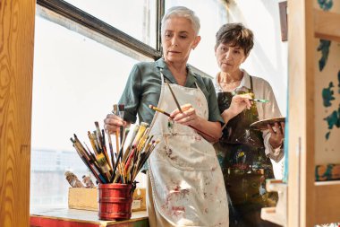 skilled female artists choosing  paintbrushes near mature woman during master class in art workshop clipart