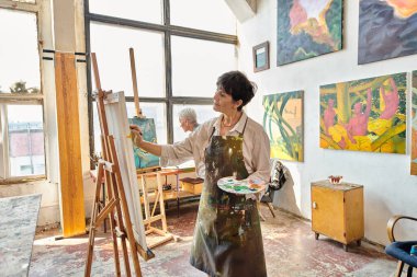 Modern ferah sanat atölyesinde, orta yaşlı bir kadın, sehpada resim yapıyor.