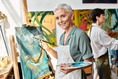 smiling mature woman painting on easel and smiling at camera in art workshop, creative process clipart