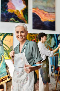 inspired and joyous mature woman with palette smiling at camera near female friend in art workshop clipart