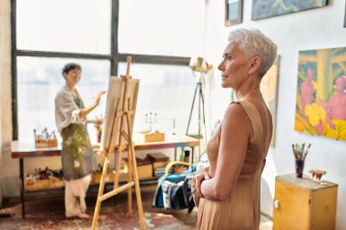 Sanat atölyesinde sehpada acı çeken kadın ressamın yanında poz veren şık, olgun model, yaratıcı süreç.