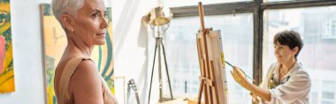 Güzel olgun model, sanat atölyesinde profesyonel kadın ressama poz veriyor, yatay afiş.