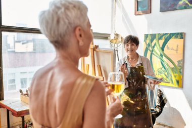 Mutlu olgun ressam, sanat atölyesinde şarap kadehiyle bulanık kadın modeli çiziyor. Yaratıcı süreç.