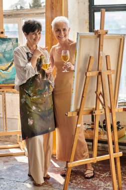 Heyecanlı kadın sanatçı ve zarif olgun model, şarap kadehleriyle stüdyoda resim sehpasına bakıyor.