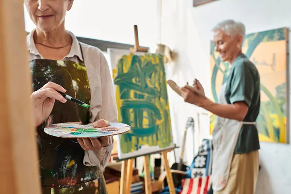 mature woman with palette near smiling female friend painting on easel in art workshop, creativity