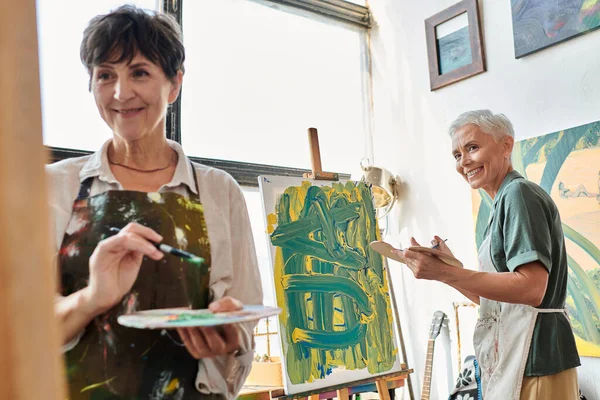 happy mature woman with palette looking at female friend painting art workshop, creative hobby