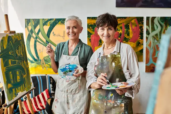 joyful mature female friends holding palettes and smiling at camera in art studio, artistic hobby