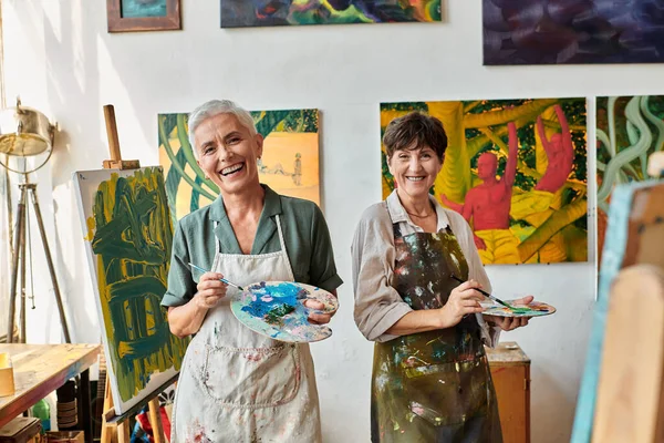 excited mature female friends holding palettes and smiling at camera in art studio, artistic hobby