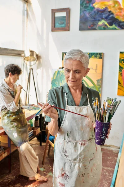 stock image thoughtful mature woman choosing paintbrush near female friend in art workshop, creative hobby