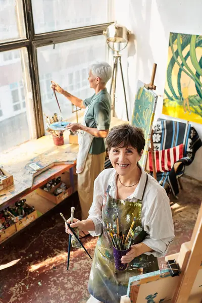 stock image happy mature woman holding paintbrushes and looking at camera near female friend in crafts workshop