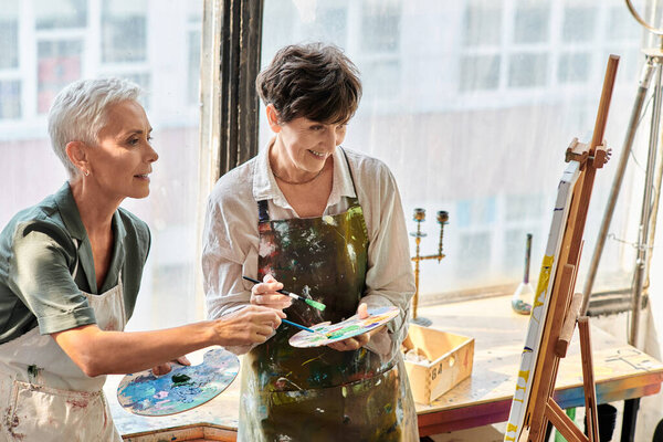 зрелые художницы смешивают цвета на палитрах возле мольберта в мастерской, мастер-класс по живописи