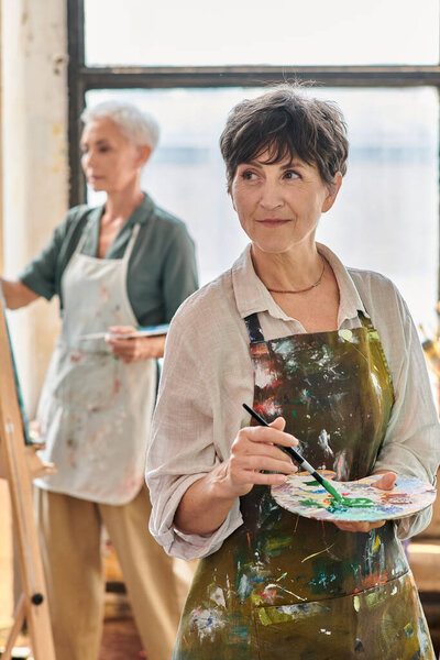 улыбающаяся зрелая женщина, держащая цветовую палитру и отворачивающаяся во время мастер-класса в художественной мастерской