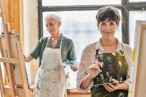 веселая зрелая женщина, держащая цветовую палитру и улыбающаяся перед камерой во время мастер-класса в художественной студии