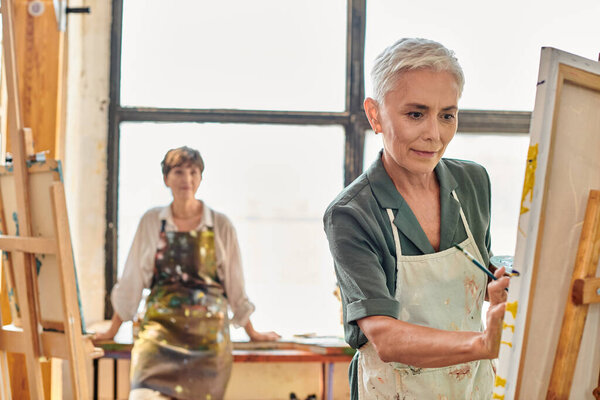 концентрированная зрелая женщина в фартуке на мольберте на мастер-классе в художественной мастерской, творческое хобби