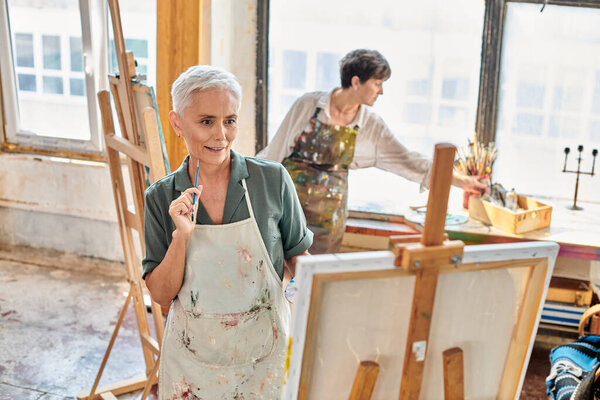 вдумчивая зрелая женщина смотрит на мольберта рядом с квалифицированной художницей в мастерской, мастер-класс