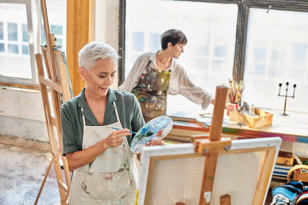 улыбающаяся зрелая женщина смешивает краски на цветовой палитре рядом с опытным художником в мастерской, мастер-класс