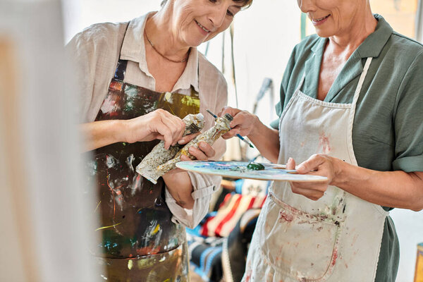 улыбающиеся зрелые женщины, смешивающие краски на цветовой палитре во время мастер-класса живописи в художественной студии