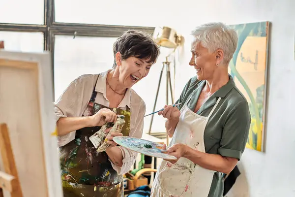 excited mature woman laughing near female friend while mixing paints on color palette, master class