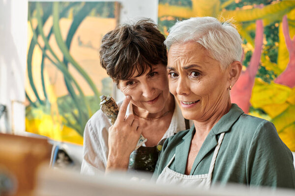 положительные и задумчивые зрелые женщины смотрят на мольберт во время мастер-класса живописи в художественной мастерской