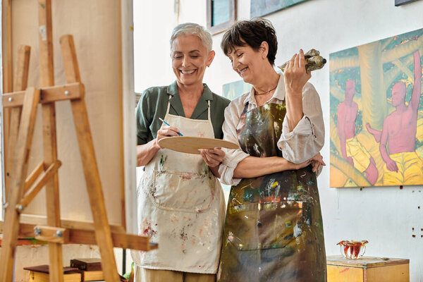 веселая зрелая женщина смешивает краску на палитре рядом с улыбающейся художницей, мастер-класс в художественной студии