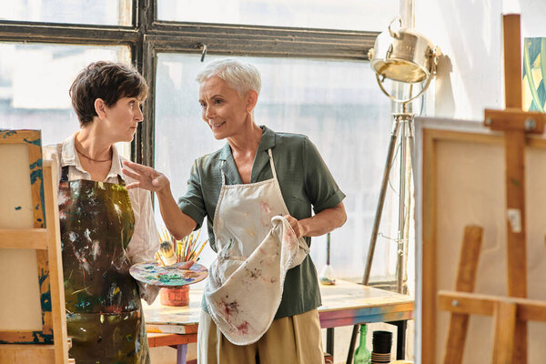 позитивная художница разговаривает со зрелой женщиной в фартуке возле мольберта во время мастер-класса в художественной студии