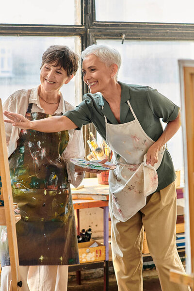 улыбающаяся художница, указывающая на мольберта рядом с счастливой зрелой женщиной во время мастер-класса в художественной студии