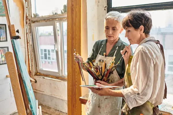 skilled female painter helping mature woman to choose paintbrush during master class in art workshop
