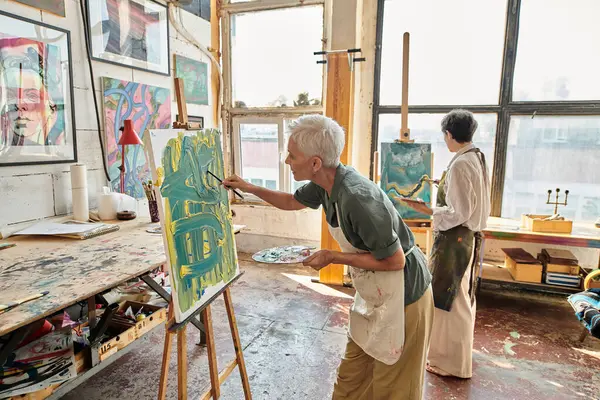 talented mature women painting on easels in modern art workshop with colorful pictures on walls