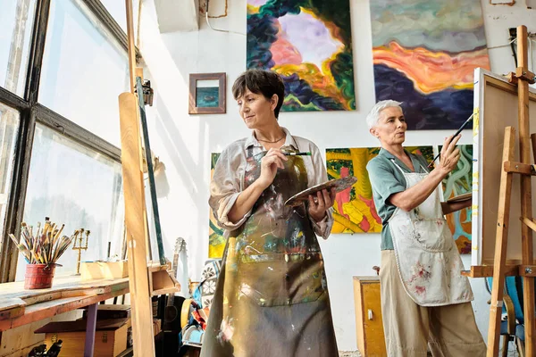 creative mature female artists in aprons pointing on easels in art studio, creative hobby