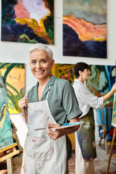 stock image inspired and joyous mature woman with palette smiling at camera near female friend in art workshop