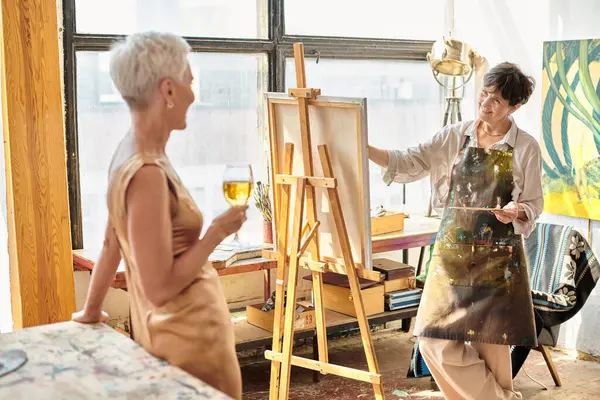 joyful mature woman painting elegant female friend posing with wine glass in art studio, creativity