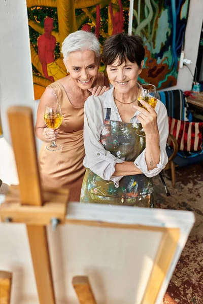 delighted mature female friends with wine glasses looking at easel in art studio, artist and model