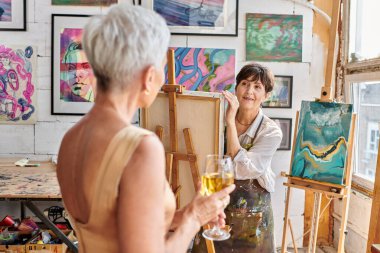 Sanat atölyesinde şarap kadehiyle poz veren bulanık orta yaşlı bir modele bakan esinlenmiş kadın sanatçı.
